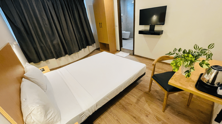 Sanctum Suites, Richmond Road, Bangalore offers a room with a double bed, a wooden desk with a chair, black curtains, and a wall mounted television.