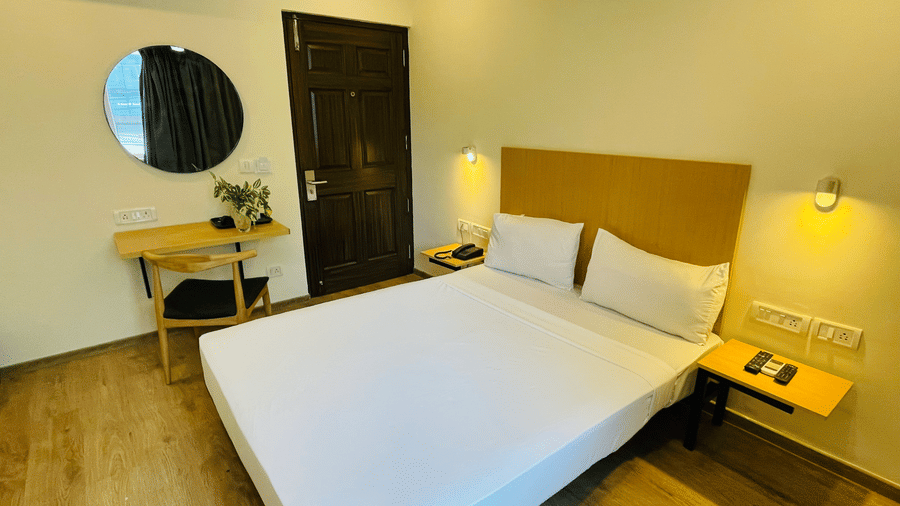 A room at Sanctum Suites, Richmond Road, Bangalore features a double bed with white linen, a wooden desk with a round mirror, a wall mounted A/C, and a dark wood door.