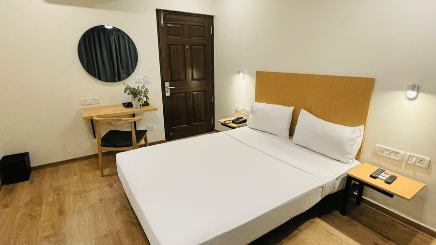 A bedroom at Sanctum Suites, Richmond Road, Bangalore features a double bed, a wall mounted A/C, a small desk with a chair, and wood effect flooring.