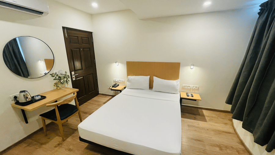 Sanctum Suites, Richmond Road, Bangalore provides a room with a single bed, a wooden desk with a kettle, a round mirror, and a wall mounted A/C.