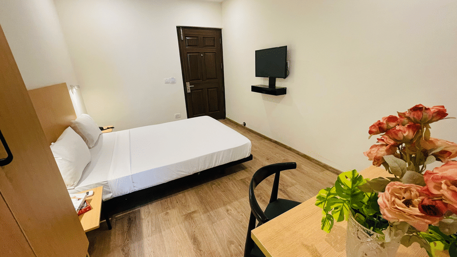 A bedroom at Sanctum Suites, Richmond Road, Bangalore featuring a double bed, a wooden desk with flowers, a wall mounted television, a chair, wardrobe, and a dark wood door.