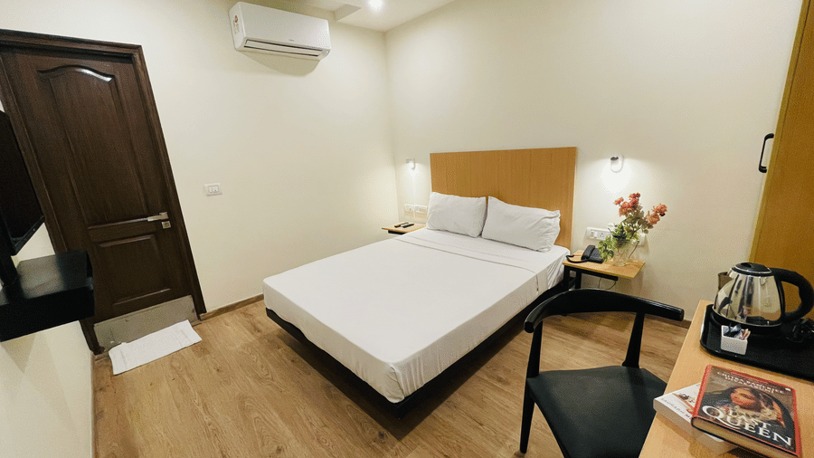 A bedroom at Sanctum Suites, Richmond Road, Bangalore features a double bed, a television, a desk with a kettle, a chair, and a dark wood entrance door.