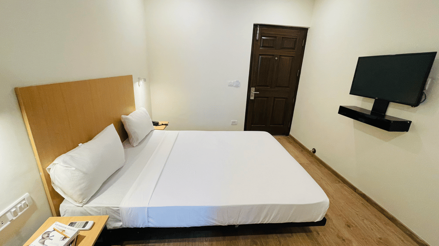 A bedroom at Sanctum Suites, Richmond Road, Bangalore with a double bed, a wall mounted television, a dark wood door, and a bedside tables.