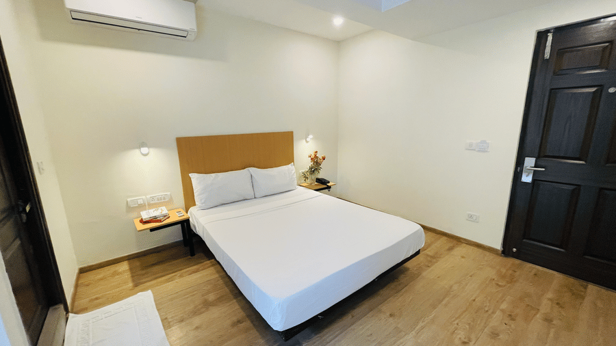 A bedroom at Sanctum Suites, Richmond Road, Bangalore featuring a double bed, a dark wood door, an air-conditioning unit, and wood effect flooring.