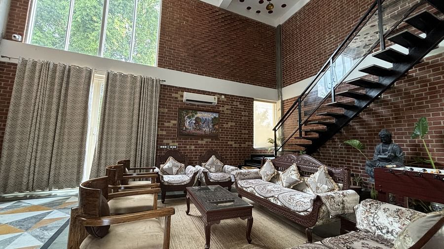 A living room at Tisya Farms, Gurgaon with multiple sofas, large windows, brick walls, and an overhead staircase.