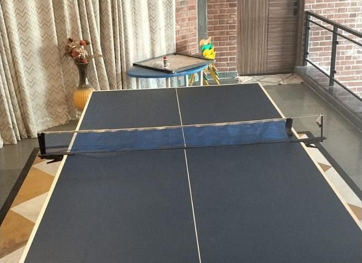 A table tennis setup indoors, featuring a blue table, a net, and paddles at Tisya Farms, Gurgaon.