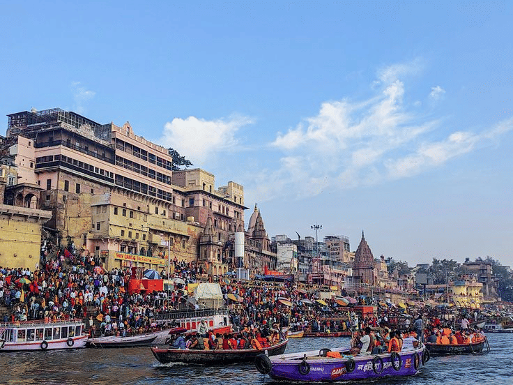 A view from the water showing boats on the river and buildings along the stone ghats under a blue sky | HHI Varanasi