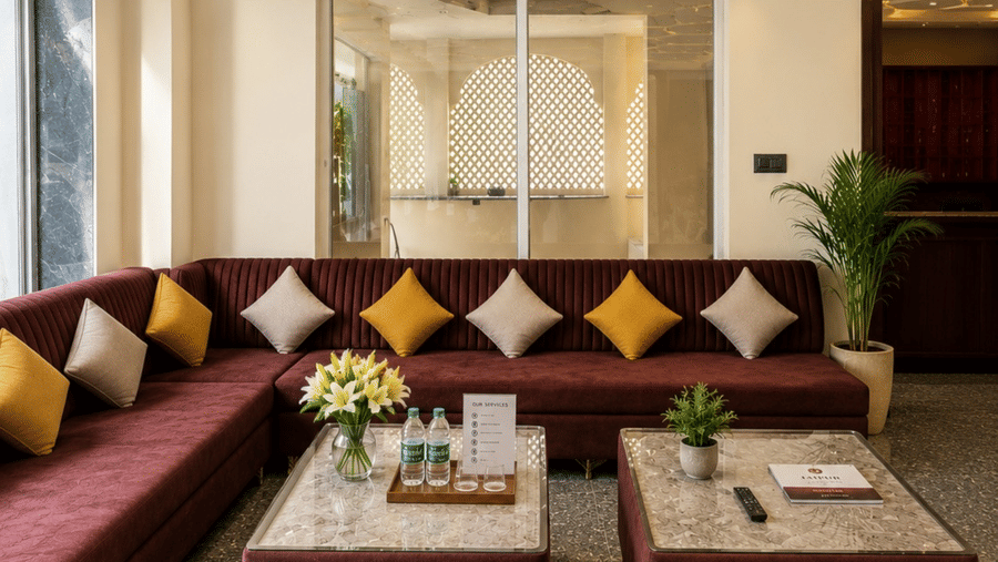 A modern hotel lounge area featuring a long, maroon L-shaped sectional sofa decorated with alternating yellow and white throw pillows, two low rectangular coffee tables on a speckled tiled floor, and a geometric honeycomb-patterned ceiling with recessed li