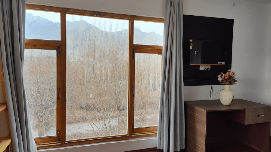 Large wooden-framed windows in a  bedroom showing a serene view of distant mountains at Indus Serenity Retreat.
