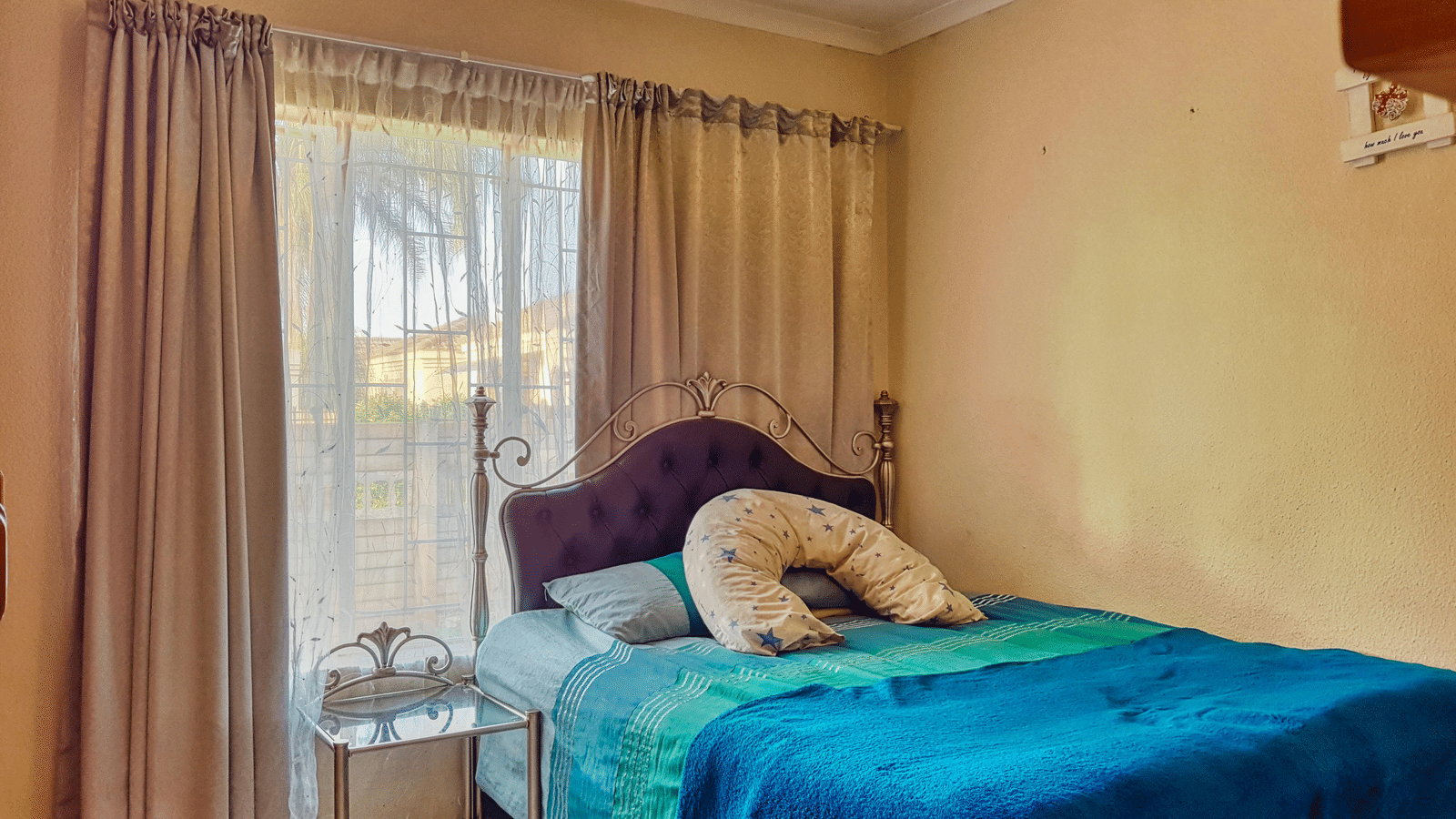 A bedroom featuring a double bed with blue sheets spread neatly over it with beige curtains covering the window behind it.