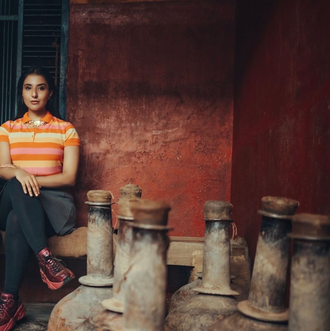 A woman in a vibrant striped polo shirt and maroon sneakers sits poised against a rustic red wall, surrounded by large, weathered clay vessels in an earthy, traditional setting - Kastoor x Sawai Hospitality