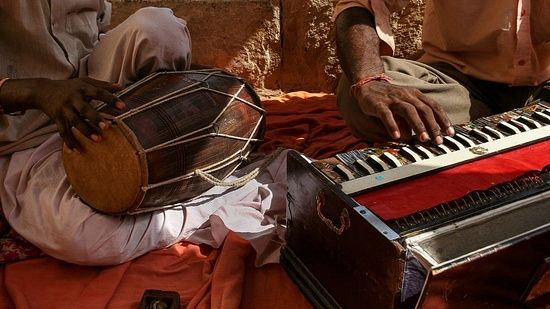 A man sitting on the floor with a dholak in his lap and a harmonium beside him, immersed in music