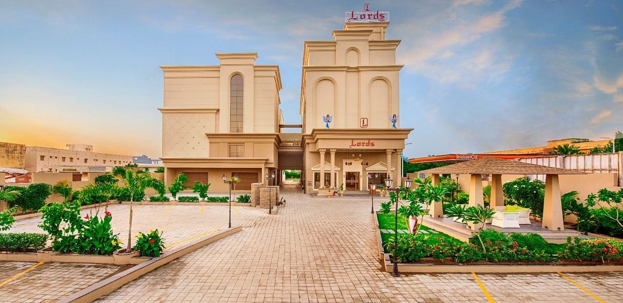 A wide shot of a light-coloured, multi-storey hotel building with a prominent entrance and surrounding greenery, including paved walkways and some parked cars, under a partly cloudy sky at either dawn or dusk at Lords Inn Motikhavdi, Jamnagar.