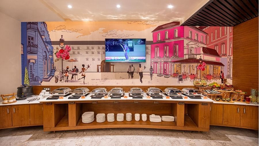 A buffet spread is presented on a long wooden counter with various covered dishes and white containers, set against a backdrop of a colourful mural depicting a European street scene and a television screen at Lords Inn Motikhavdi, Jamnagar.