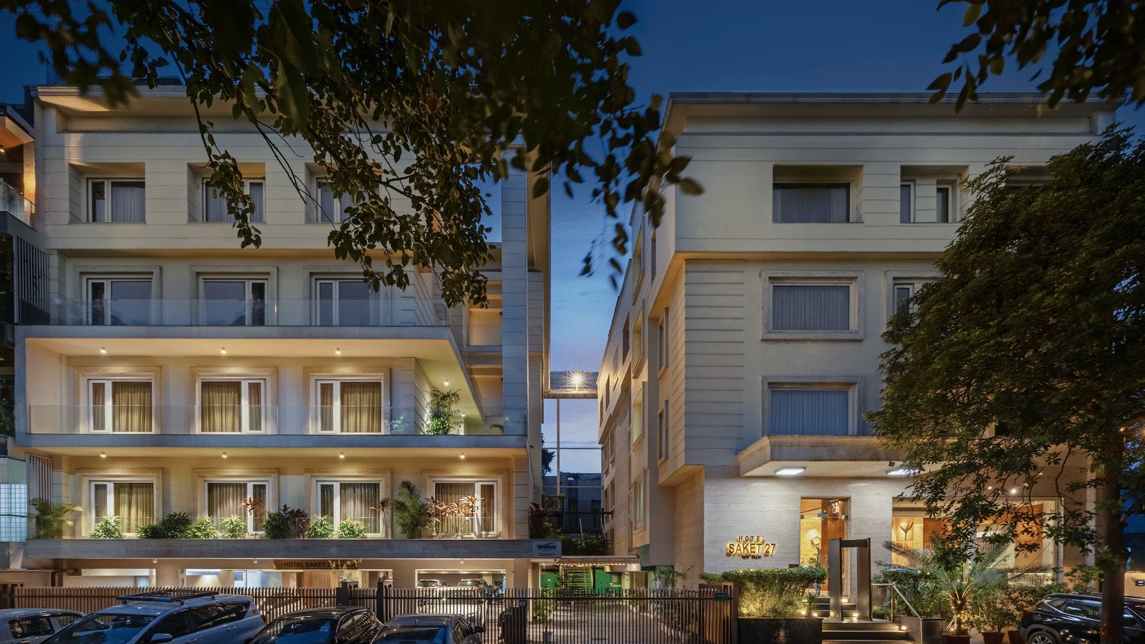 Night view of Hotel Saket 27’s exterior showcasing a multi-story building with balconies, illuminated façade, a grand entrance, and an inviting ambience for visitors.