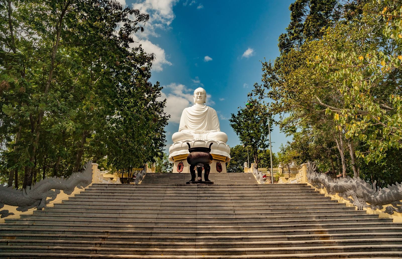 Statue at the top of a wide staircase, framed by trees against a bright sky.