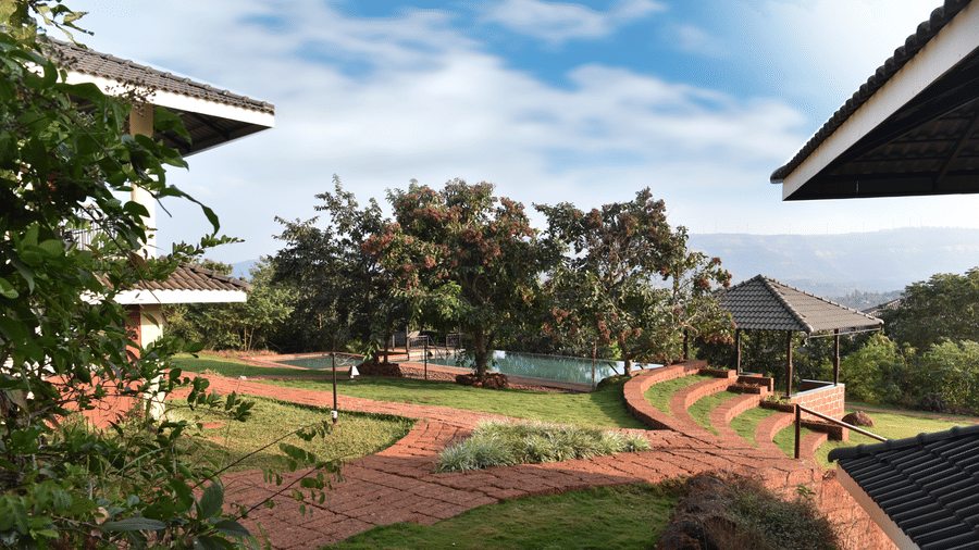 Forest Escape, an eco-friendly resort near Pune, featuring a wider view of the resort grounds with gardens and trees under a partly cloudy sky