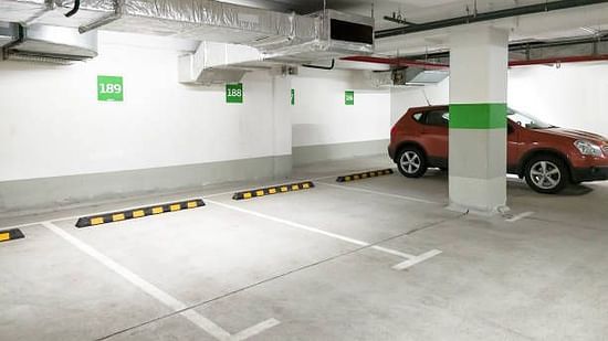 An indoor parking garage with a red car parked inside. White lines mark parking spaces on the gray concrete floor. Overhead pipes and a support column are visible.