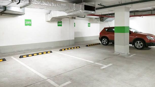 An indoor parking garage with a red car parked inside. White lines mark parking spaces on the gray concrete floor. Overhead pipes and a support column are visible.