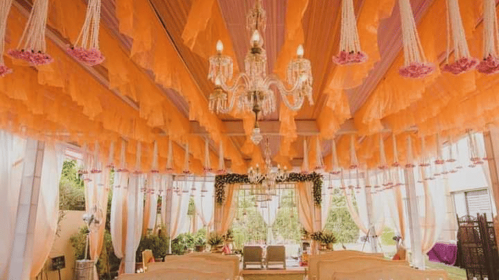 A brightly decorated hall with orange fabric ceiling drapes, elegant chandeliers, and rows of gold-framed chairs at Golden Tulip Kukas, Jaipur.