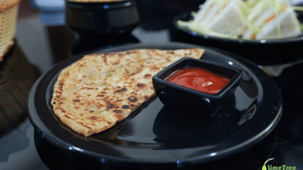 A freshly cooked flatbread served on a black plate with a small bowl of red dipping sauce, with sandwiches in the background.