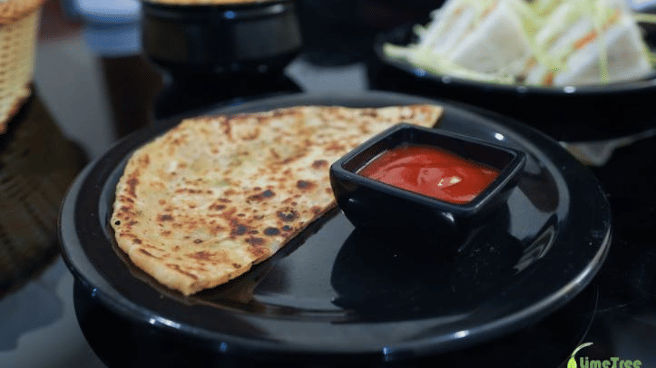 A freshly cooked flatbread served on a black plate with a small bowl of red dipping sauce, with sandwiches in the background.