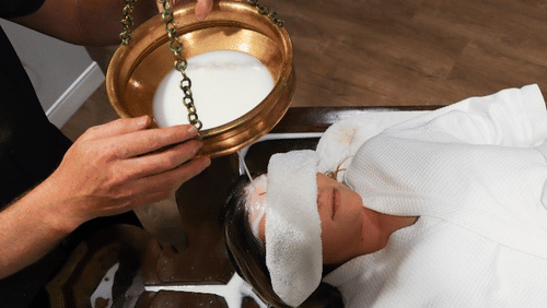 A person receiving an Ayurvedic shirodhara treatment, with a person pouring a liquid onto their forehead from a bowl at YO1 Longevity & Health Resorts, Catskills