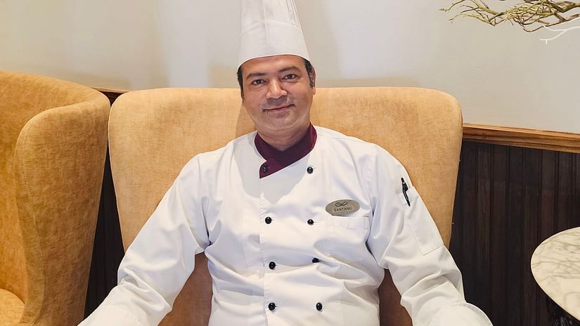 A professional chef in a white uniform and toque sitting in a cozy restaurant setting with warm lighting and elegant decor.