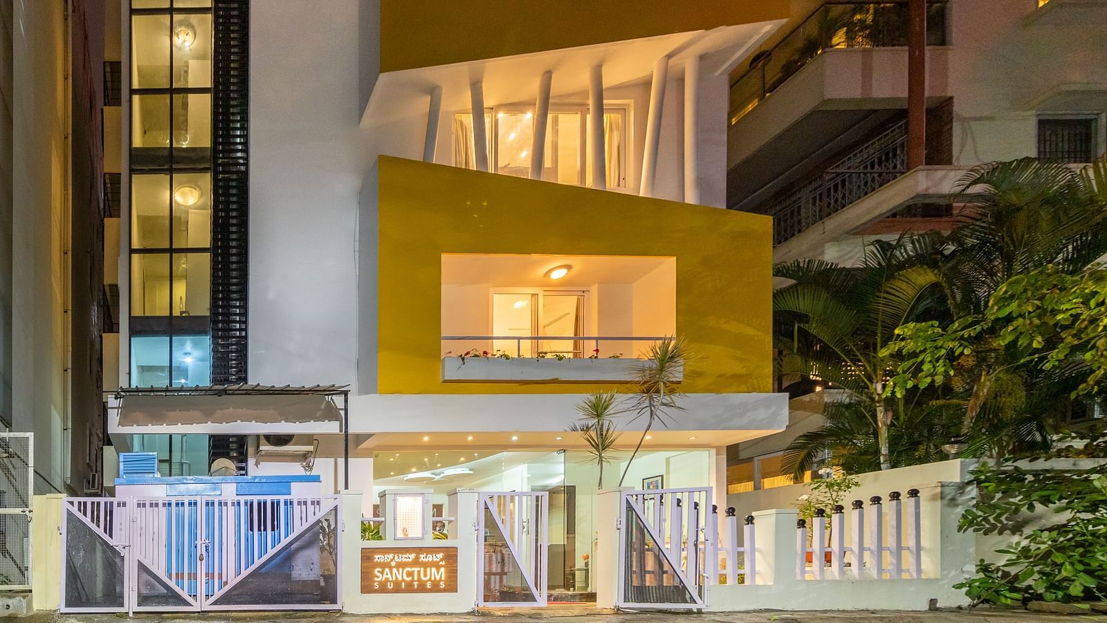A facade of Sanctum Suites Domlur, Bangalore with warm lighting and view of balconies shot at night.