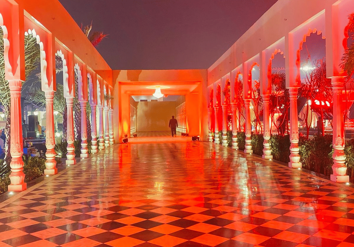 A long, arched walkway within a palace or courtyard, illuminated with a vibrant red-orange light that reflects strongly off the checkered floor tiles at night.