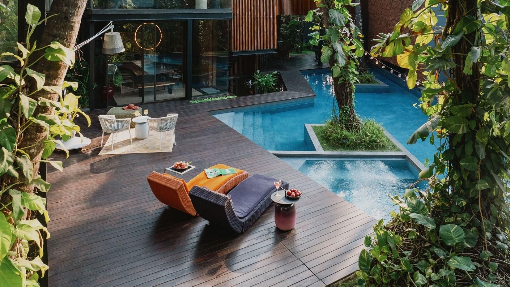 An image of a swimming pool surrounded by plants and on the side of of a pool there are lounger beds