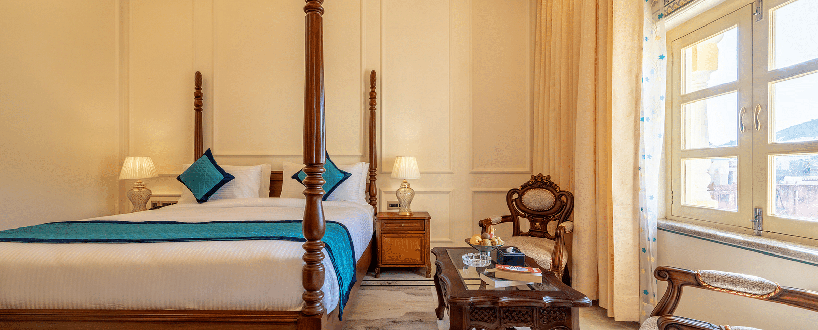 a bedroom featuring a king size bed along with two arm chairs placed in front of a coffee table with a decorative wooden frame behind it - Brij Castle, Jaipur