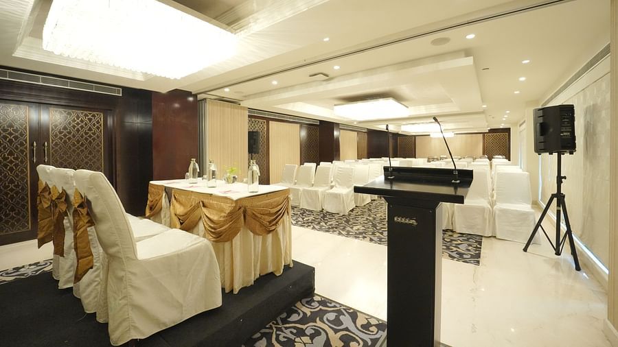 A conference room setup with a podium, speaker system, and chairs with white covers and gold sashes - Fragrant Nature Cochin