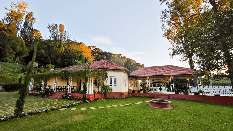 A white single-story building with a red roof set on a green lawn with tall trees against a bright blue sky.