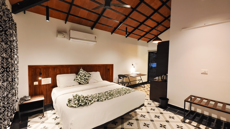 The well-lit premium room at Ibex Resorts, Malampuzha (Kava Eco Camp and Caravan Park), featuring a large bed, bedspreads, pillows, tables, a chair, and a wall mounted air-conditioner.