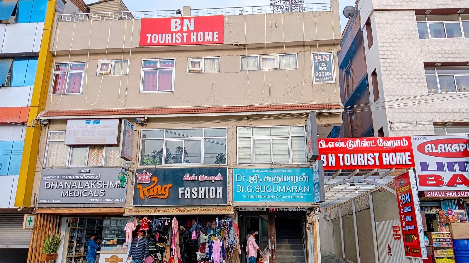 A three-story building in Ooty, India, features BN Tourist Home signage. Street-level shops include Dhanalakshmi Medicals and King Fashion. A doctors office is also visible.