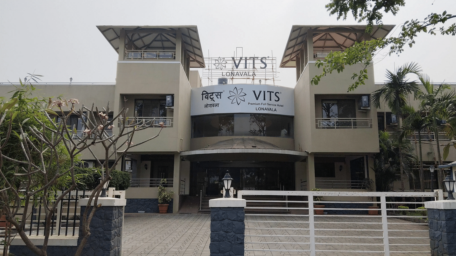 Front view of a gated hotel building with 'VITS' signage, twin tower structures, and a central entrance flanked by greenery and paved driveway.