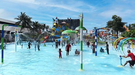 Children and adults in a shallow splash pool with water jets, spray arches and a mushroom fountain, with slides in the background at MGM Dizzee World, Chennai.