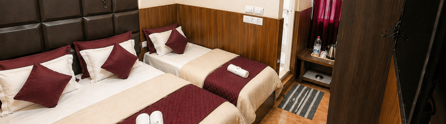 A double bed and a single bed arranged together with folded towels, a wardrobe, and a television in the Deluxe Triple Room at BN Tourist Home, Ooty.