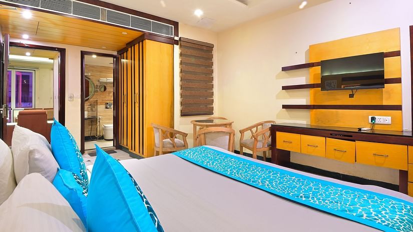 Room at Hotel Hari Piorko in Paharganj featuring a plush bed facing the TV mounted on the wall along with a counter next to which a sitting space is arranged with a round table and four comfy chairs as the doorway to the bathroom is right beside it