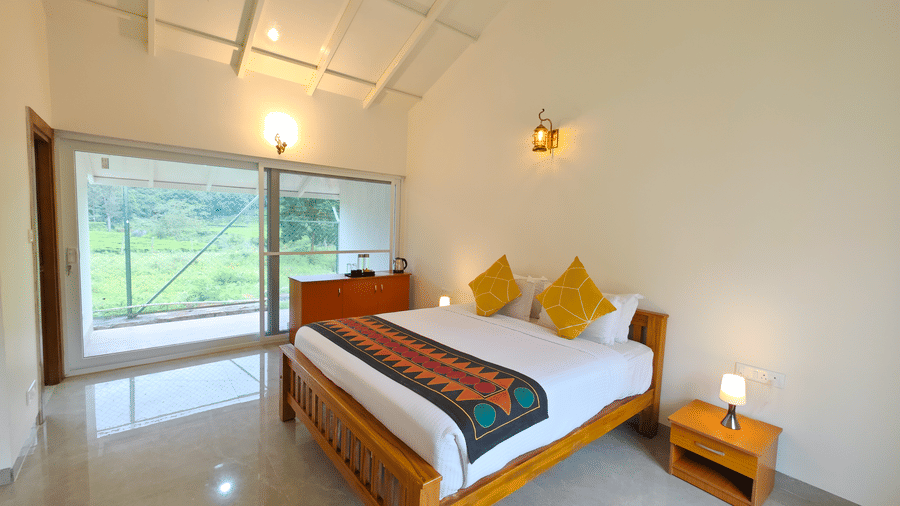 Tiger room at Ibex Resort, Kotagiri, shows a large bed on a stone floor under a beamed ceiling. Sliding doors to the exterior sit on one wall, with a small cabinet on another.