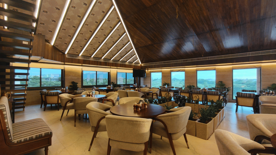 Spacious dining room at The Andhra Leaf restaurant, Ni Central Park, Mysore, featuring round dining tables, comfortable armchairs, and a slanted wooden ceiling with recessed lighting.