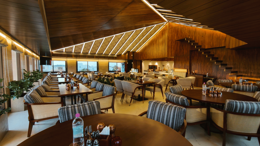 Warmly lit interior of The Andhra Leaf restaurant at Ni Central Park, Mysore, showing dark wood ceilings, round dining tables, and comfortable cushioned chairs.