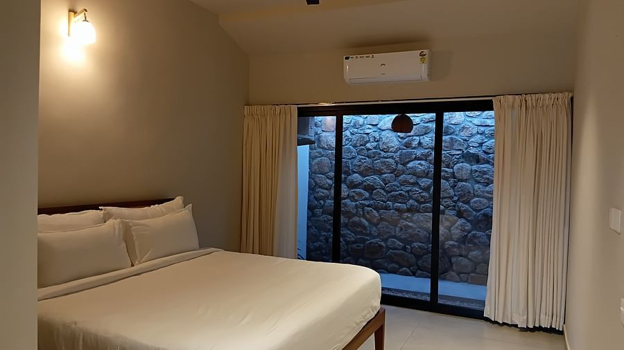A cozy bedroom at Vaasta Vagamon with a neatly made bed, a ceiling fan, and a large window overlooking a stone wall.
