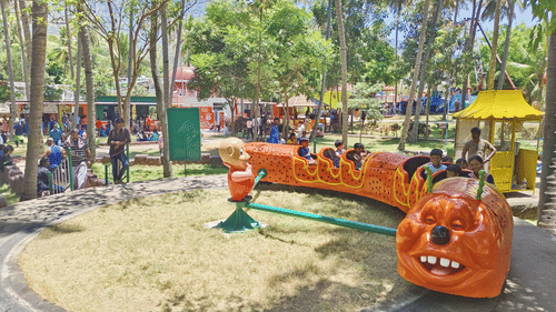 Children riding a colourful caterpillar-themed train at an outdoor amusement park, surrounded by trees and families enjoying the day.