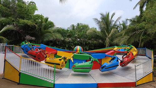 A brightly coloured amusement park ride with multiple car-like seats, set outdoors amidst lush green palm trees.