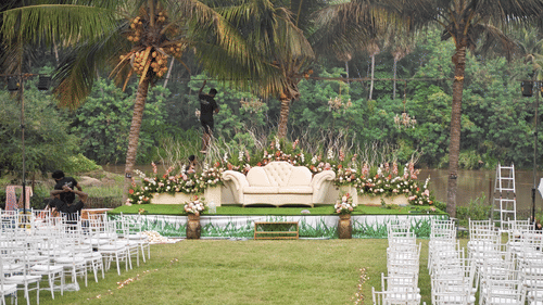 A wedding stage set against a backdrop of palm trees and a river, with rows of white chairs arranged on a lawn.