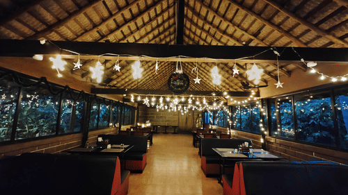 An indoor restaurant or lounge area at night, characterized by a high, textured ceiling, rows of booth seating, and numerous string lights strung across the space, creating a warm ambiance.