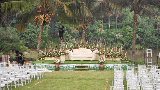 A wedding stage set against a backdrop of palm trees and a river, with rows of white chairs arranged on a lawn.