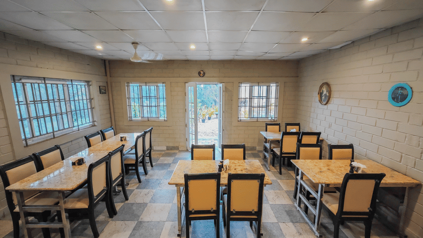 Full view of the main dining hall at Ibex Resorts Valparai Mistly showing multiple wooden tables and chairs arranged systematically on a checkered floor with bright ambient lighting and peaceful surroundings.
